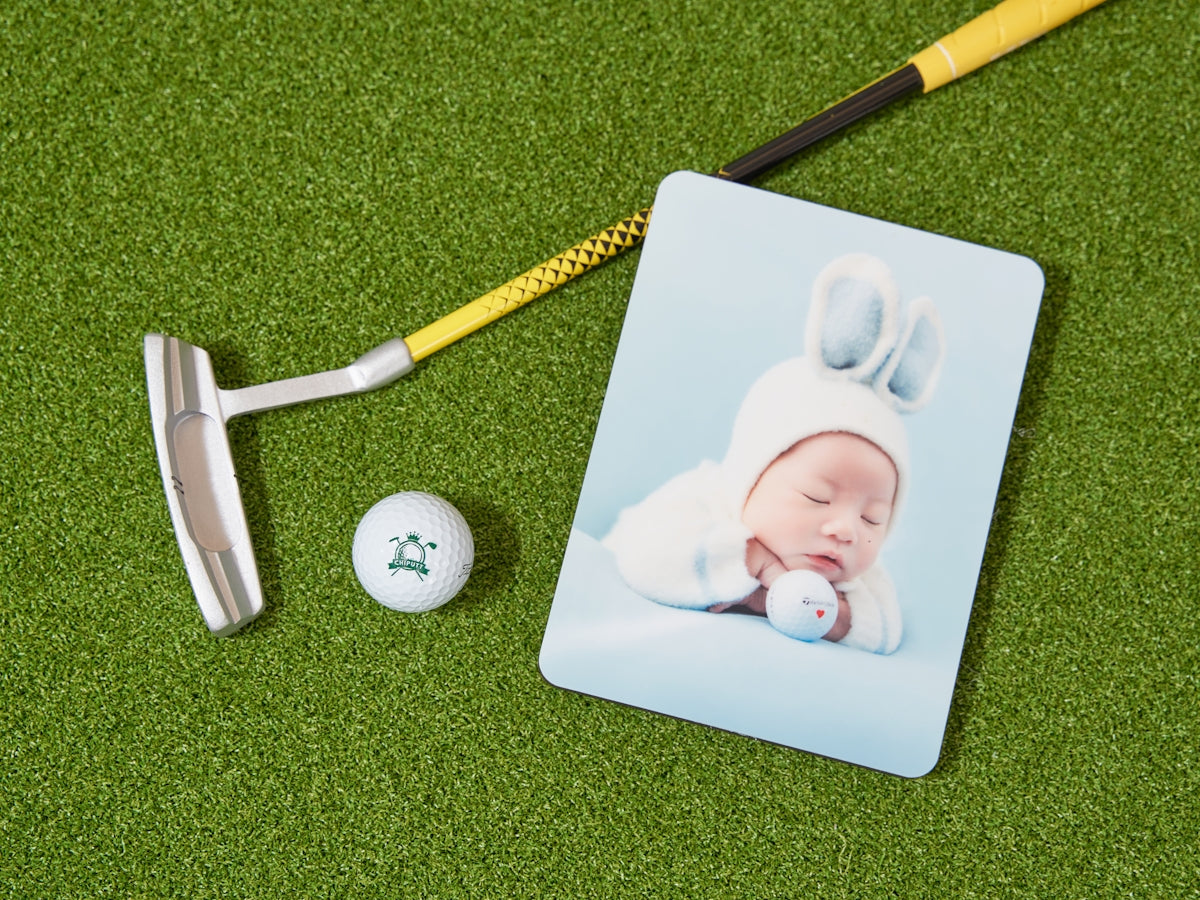 Golf putter, ball, and baby photo on green turf.